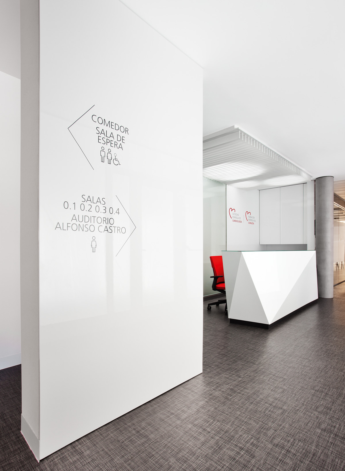 Weiß, Rot und Schwarz dominieren die Farbgestaltung im Eingangsbereich. Hinter dem geometrisch skulptural ausgebildeten Empfangstresen werden rot gepolsterte Neos-Bürostühle eingesetzt (Design: wiege).