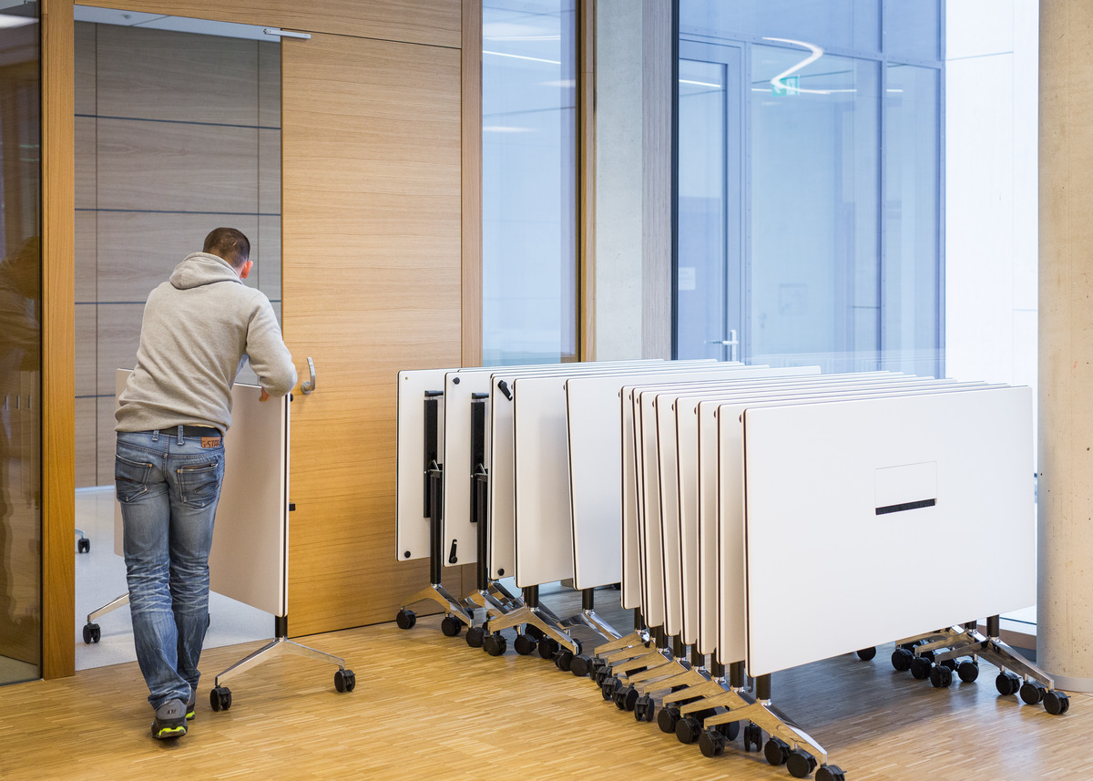 In den formschön gestalteten Tischen sind die Anschlüsse für Stromversorgung und Medientechnik fest integriert. Mit vertikal hochgeschwenkten Tischplatten lassen sie sich einfach durch das Gebäude bewegen und kompakt ineinanderstaffeln.